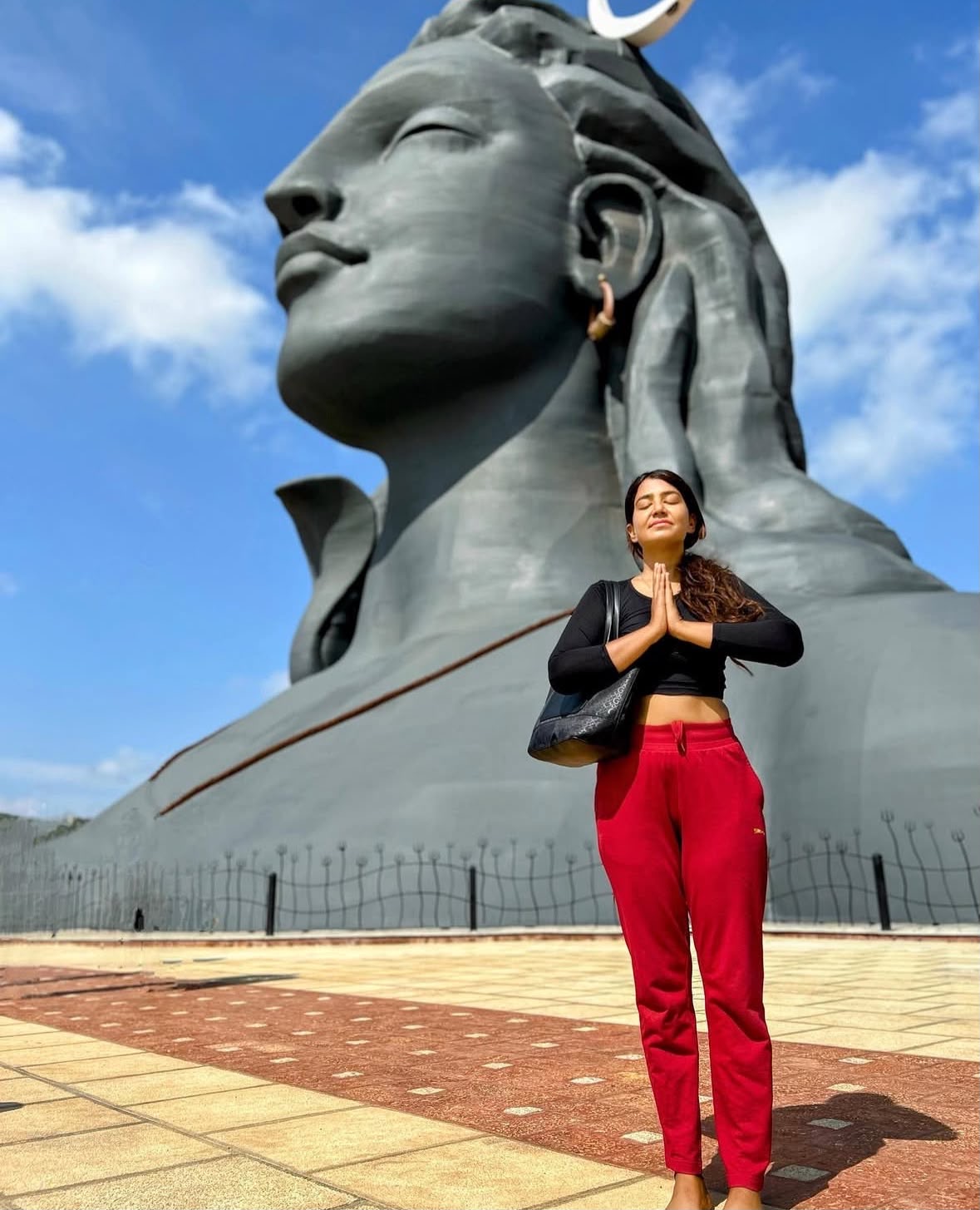 Tara standing in prayer before a monumental Shiva statue beneath a bright blue sky